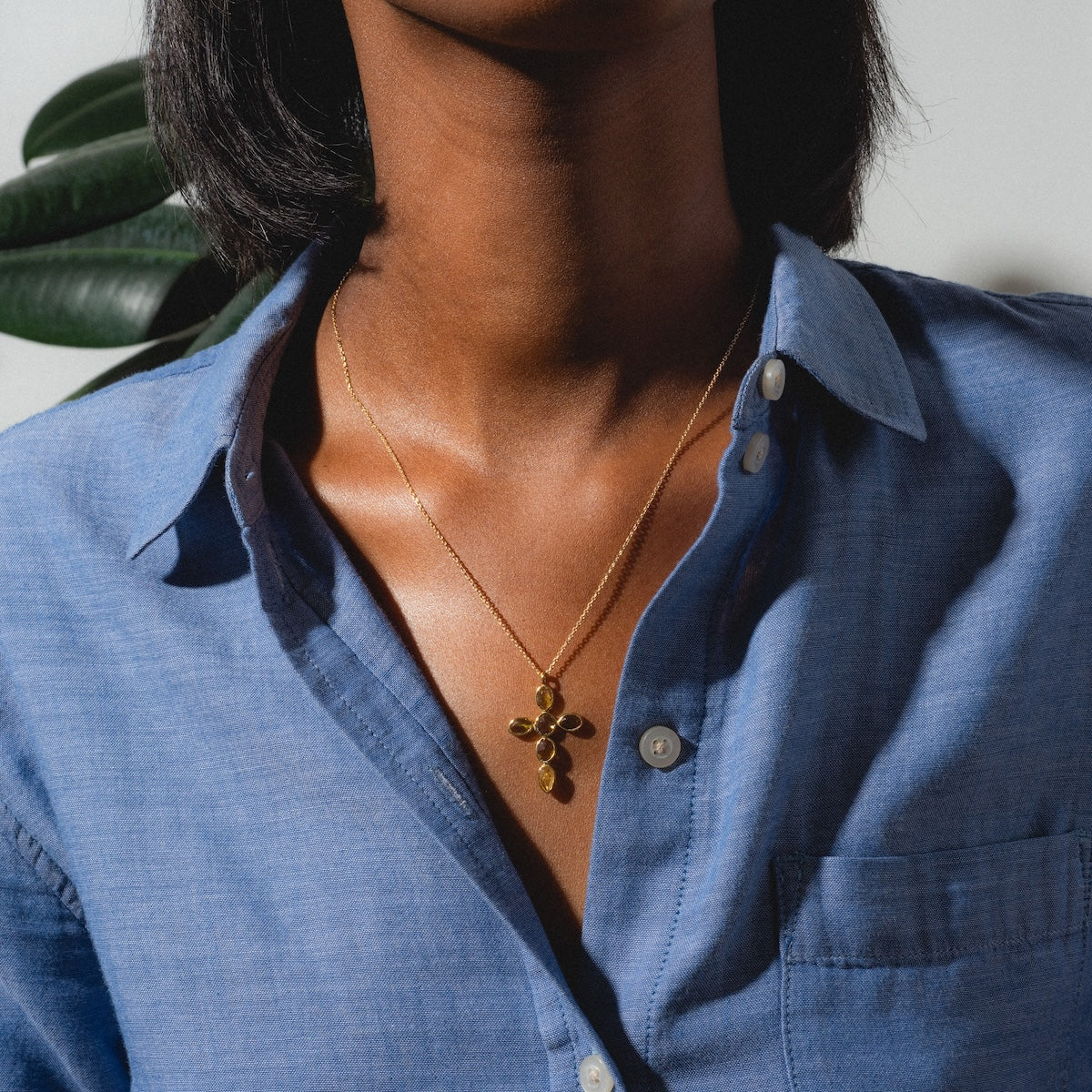 A person in a blue button-up shirt with an open collar showcases the Anika Chain Necklace—a gold necklace featuring a pendant of multiple circles—standing in soft natural light with a green plant partly visible behind them.