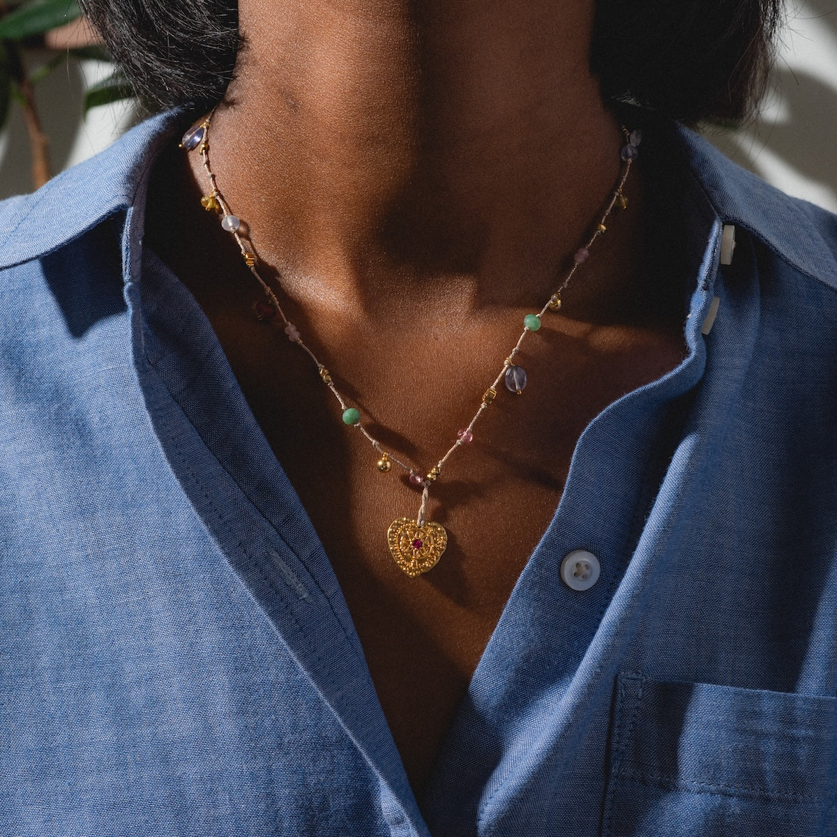 A person in a blue button-up shirt wears the Neha Necklace, a gold plated beaded necklace with a heart-shaped pendant, while standing in sunlight.