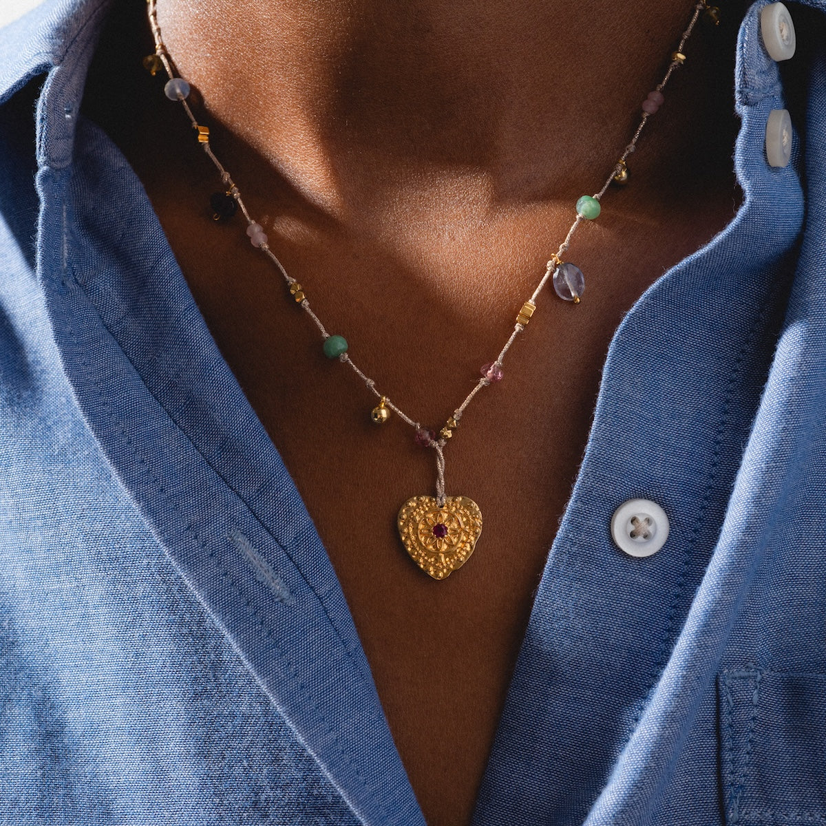 A close-up of a person wearing a blue button-up shirt and the Neha Necklace, a gold-plated necklace with a heart-shaped pendant featuring a small red gem in the center.