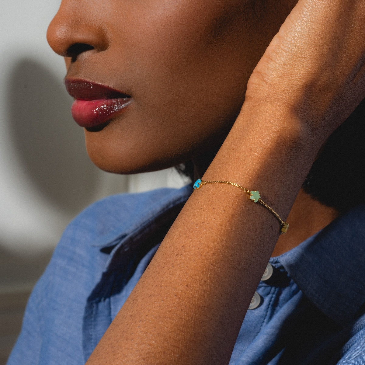 A woman with rich brown skin and glossy red lipstick wears the Star Bright Bracelet, adorned with small blue and green charms. Dressed in a blue shirt, she holds her hand near her face, highlighting the colorful accessory.