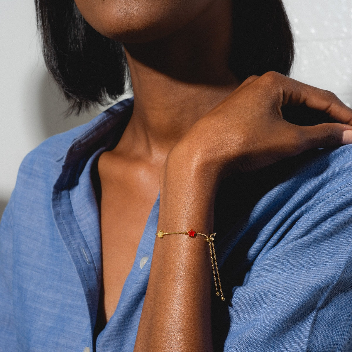 A person in a blue shirt gently rests their hand on their shoulder, featuring the Star Bright Bracelet—a delicate gold chain with a small red charm—on their wrist.