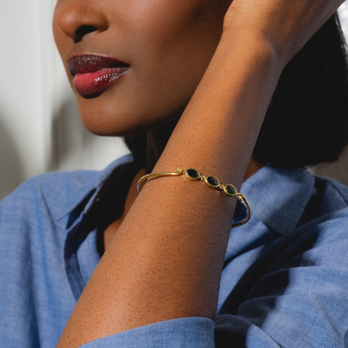 A woman in a blue shirt with glossy red lips wears the Jonc Imperial Bracelet, a gold cuff set with four dark green stones, resting her hand near her face.