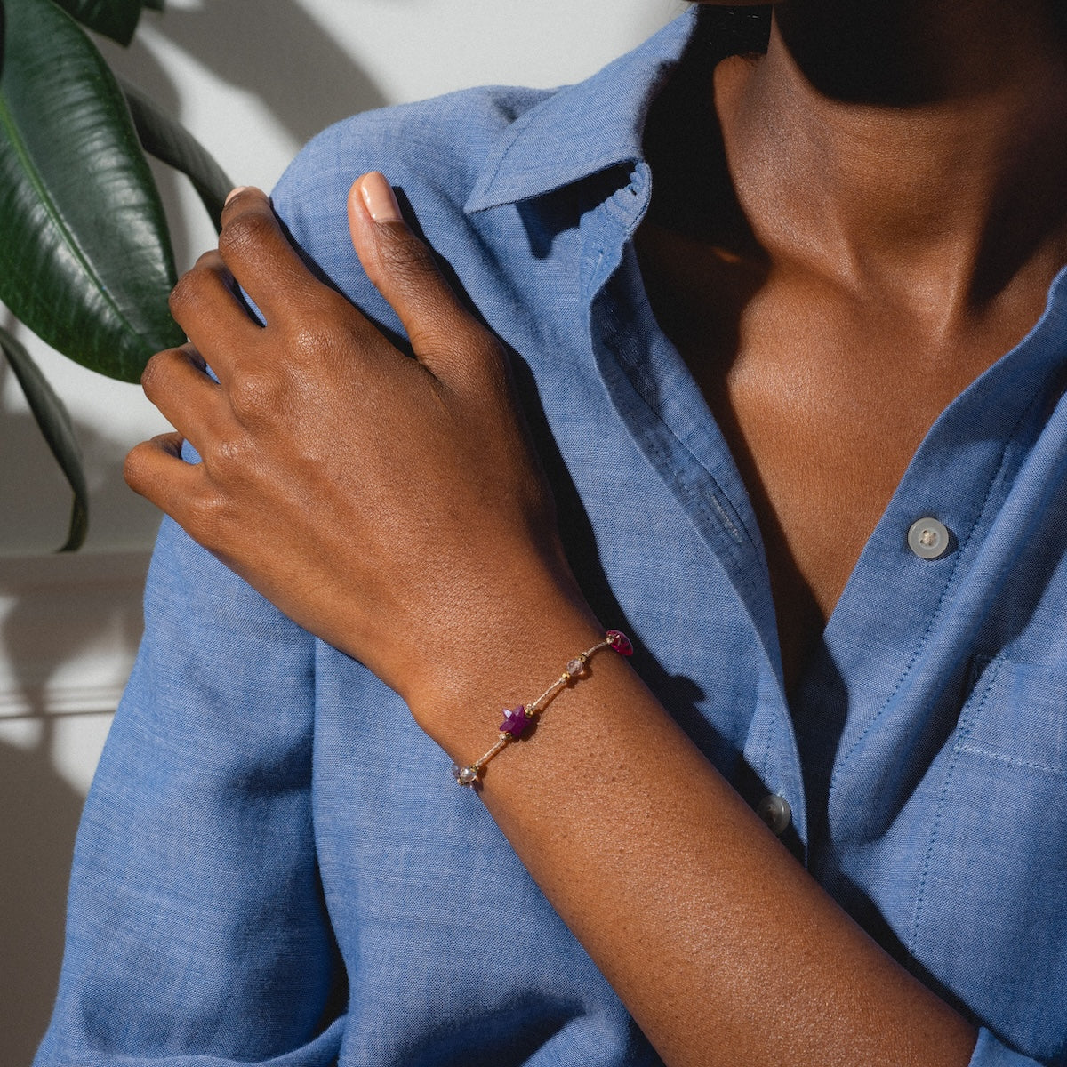 A person in a blue button-up shirt rests their hand on their shoulder, highlighting the Martha Bracelet, an elegant gold-plated piece with multi-colored tourmaline accents. A green plant leaf appears in the background.