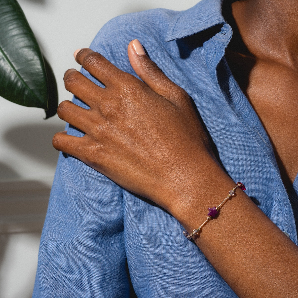 Wearing a blue button-up shirt, a person rests their hand on their shoulder, displaying the gold plated Martha Bracelet with purple gemstones on their wrist. A green leaf appears in the background.