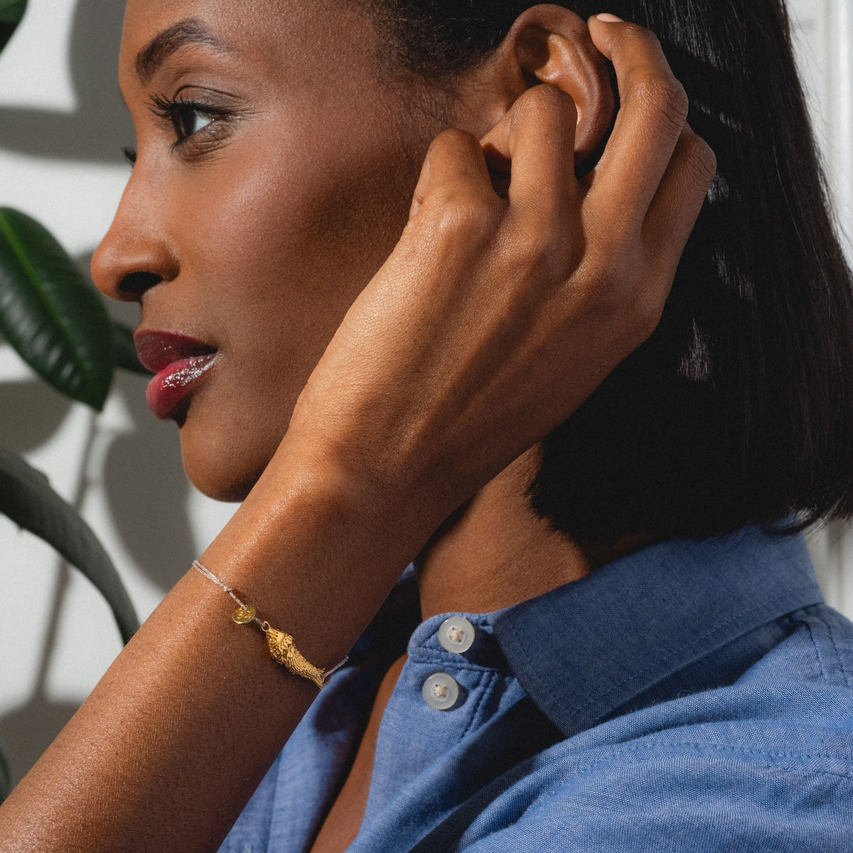 A woman in a blue collared shirt with short hair and red lipstick touches her ear, showing off the Sardina Bracelet on her wrist. She is photographed in profile with a leafy plant behind her.