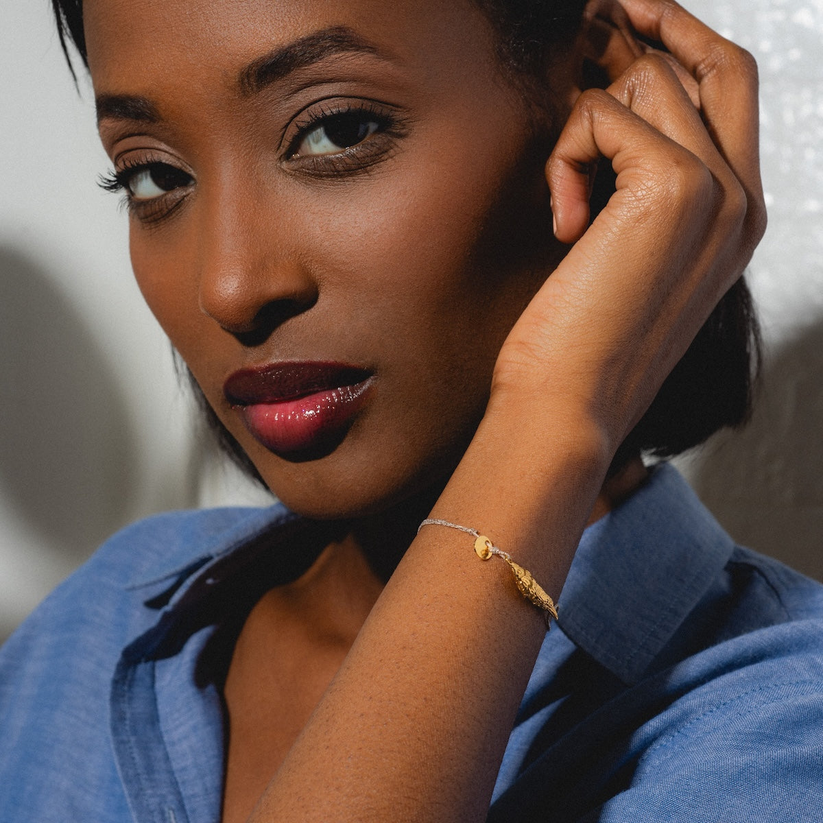 A woman with short hair and deep red lipstick, dressed in a blue shirt, poses with her hand near her ear, showcasing the Sardina Bracelet featuring a delicate gold fish charm on her wrist.
