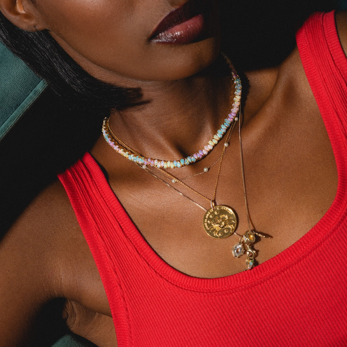 Close-up of a woman in a red sleeveless top wearing layered necklaces, including a colorful beaded choker, a gold coin pendant, and the Solid Gold Charm Clip personalized necklace. Lighting highlights her neckline and jewelry.