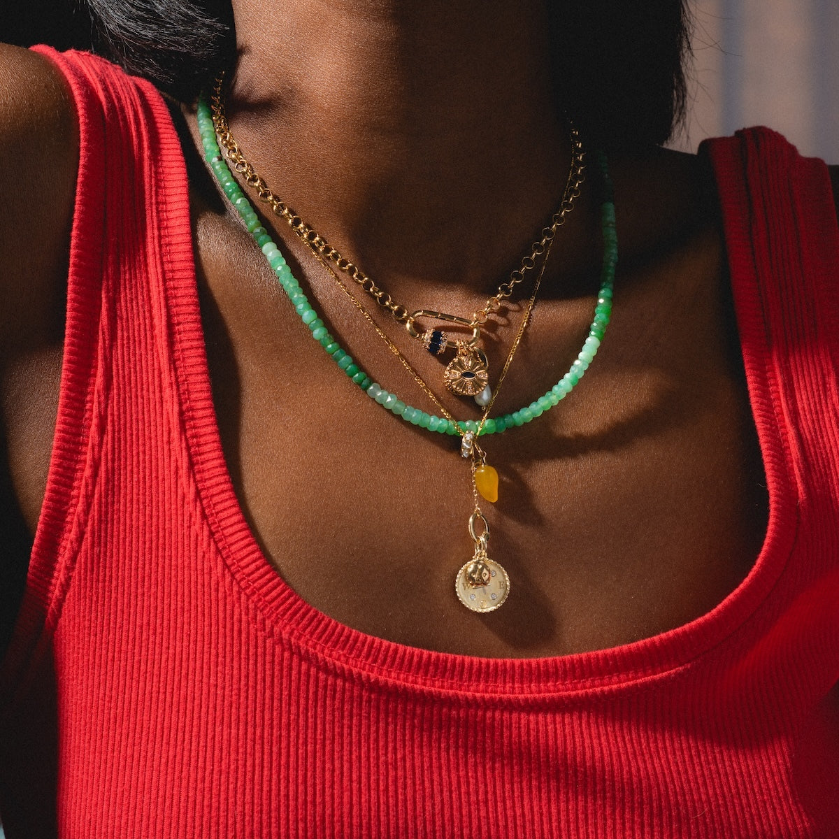 A person wears a red sleeveless top and layered necklaces, including a green beaded necklace and a gold chain with the Solid Gold Foodie Charm—shown from shoulders to collarbone.