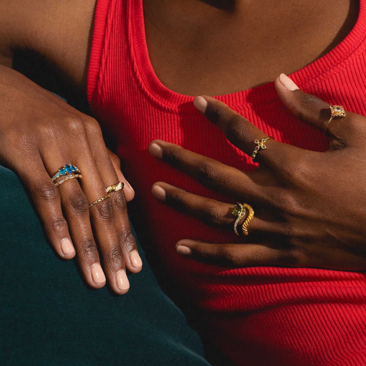 A person in a red sleeveless top displays several gold rings, including the True Colors Classic Ombre Birthstone Ring, with different designs and gemstones on their fingers, resting their hands on a dark green surface.