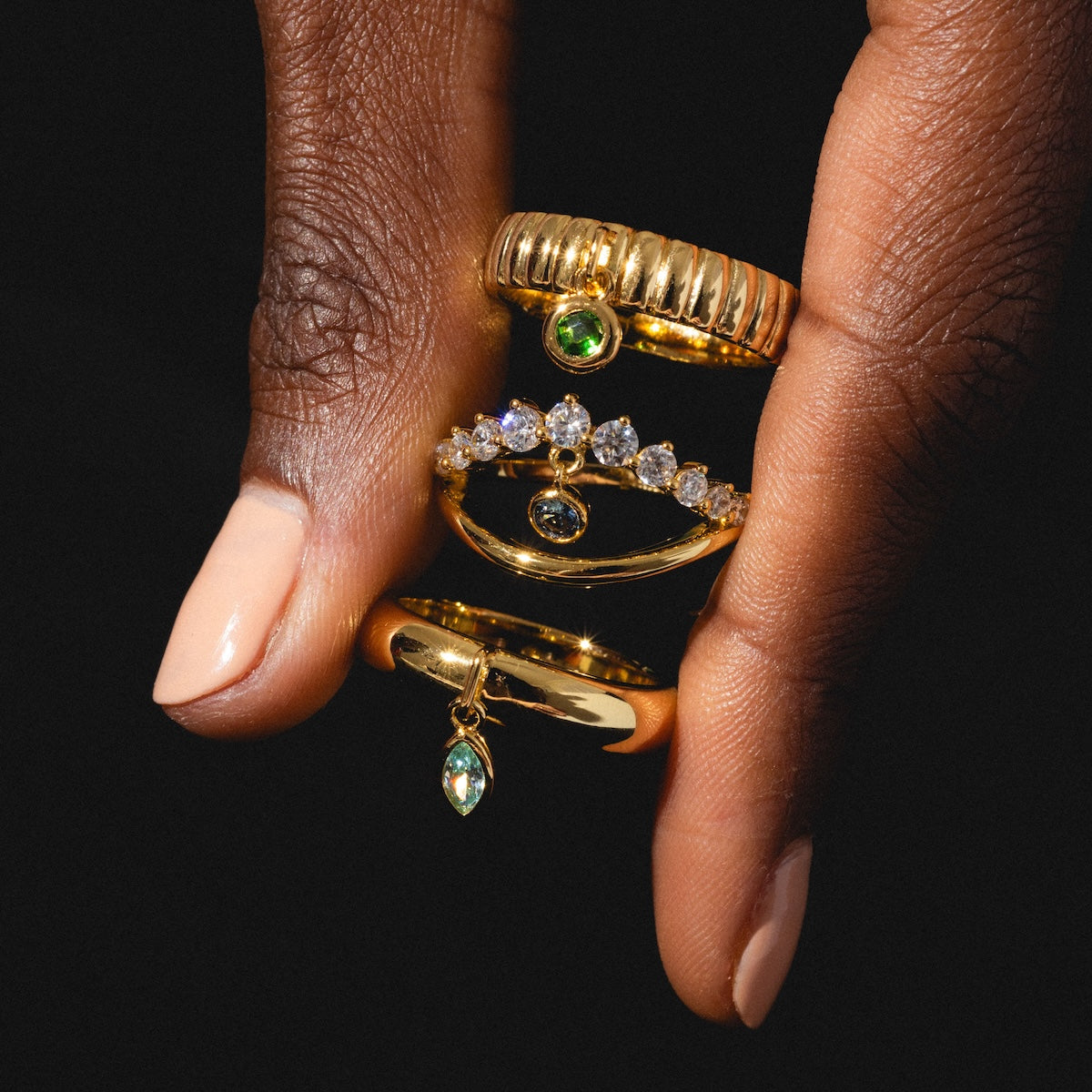 A hand with light pink nail polish displays the Mystique Evil Eye Charm Ring—14k yellow gold plated with sparkling green and clear stones—alongside gems, set against a black background.
