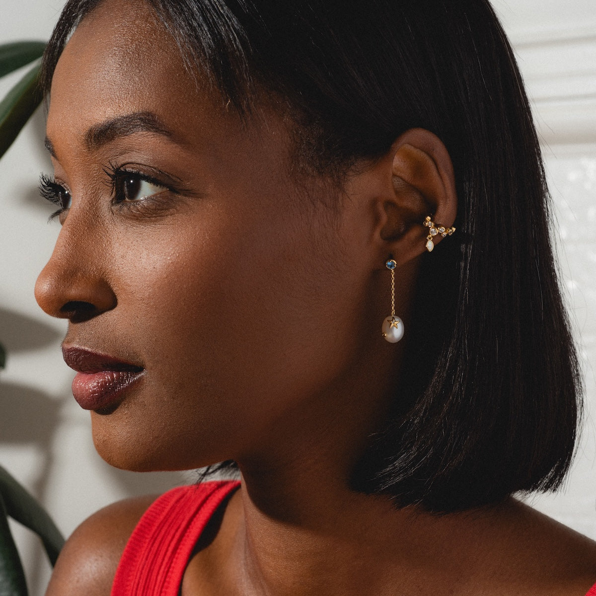 A woman with short black hair wearing a red sleeveless top, gold and pearl earrings, and the Sparkle Drop Ear Cuff gazes left, posing against a white backdrop with soft lighting.