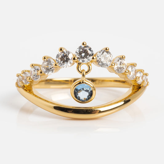 The Mystique Evil Eye Charm Ring, plated in 14k yellow gold, features a row of sparkling gems and a central dangling blue gemstone charm, shown on a white background.