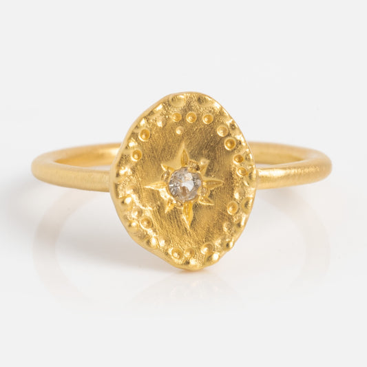 The Brahma Ring is yellow gold plated with a textured oval bezel, dotted accents, and a central white topaz in a starburst pattern, shown on a white background.