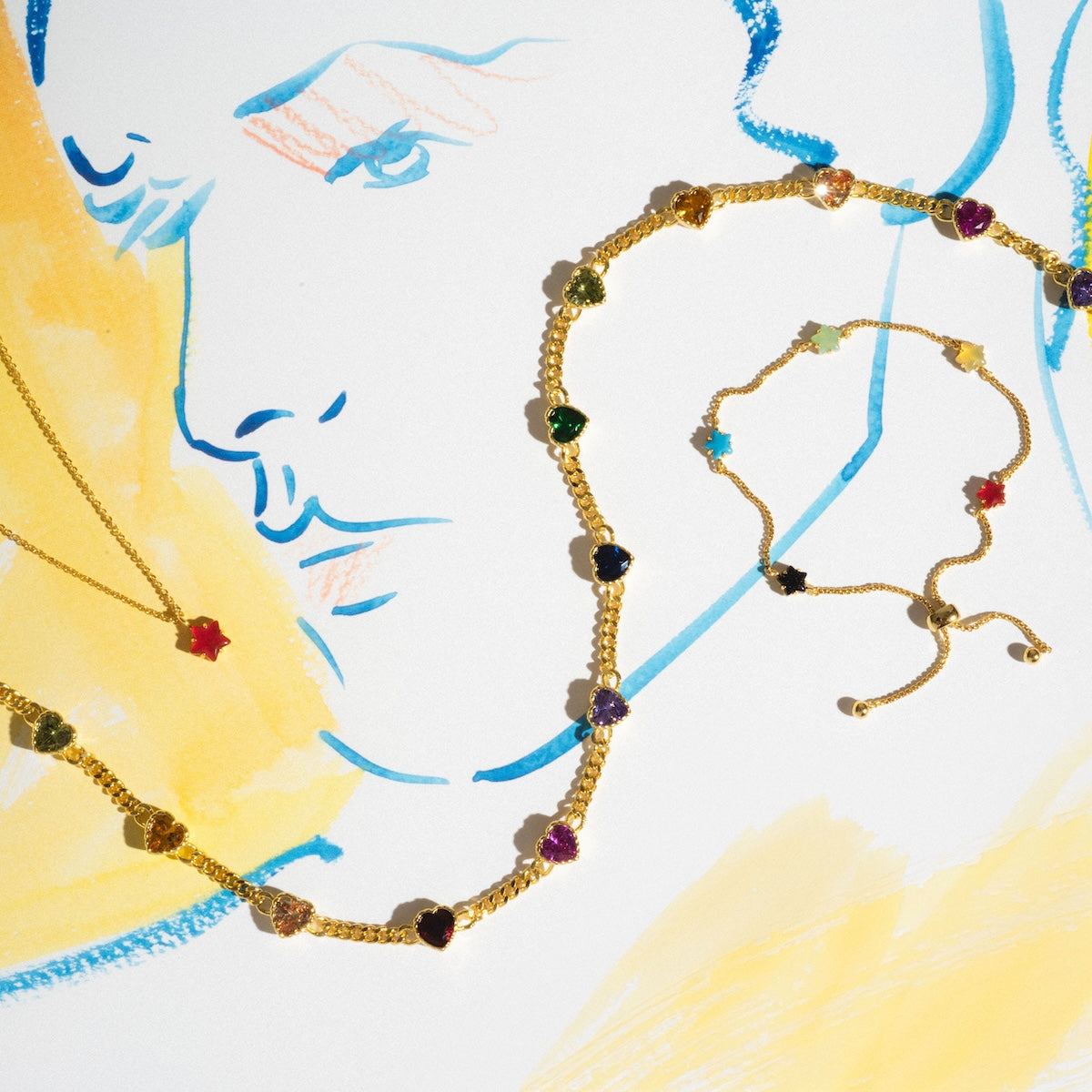 Gold jewelry with colorful gemstone charms, including a gold chain necklace and bracelet with heart-shaped stones, is displayed on an abstract blue and yellow face drawing. The Star Bright Necklace adds a playful red star to the vibrant set.