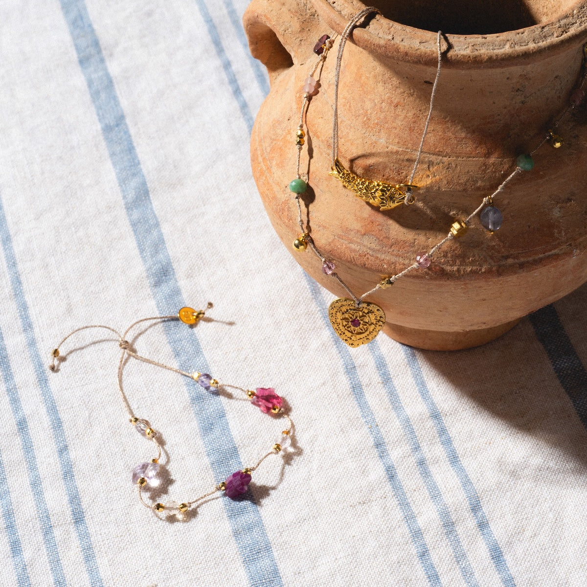 A beaded Sardina Bracelet and a gold heart pendant necklace by Sophie Deschamps Bijoux are displayed on striped fabric beside a rustic clay pot.