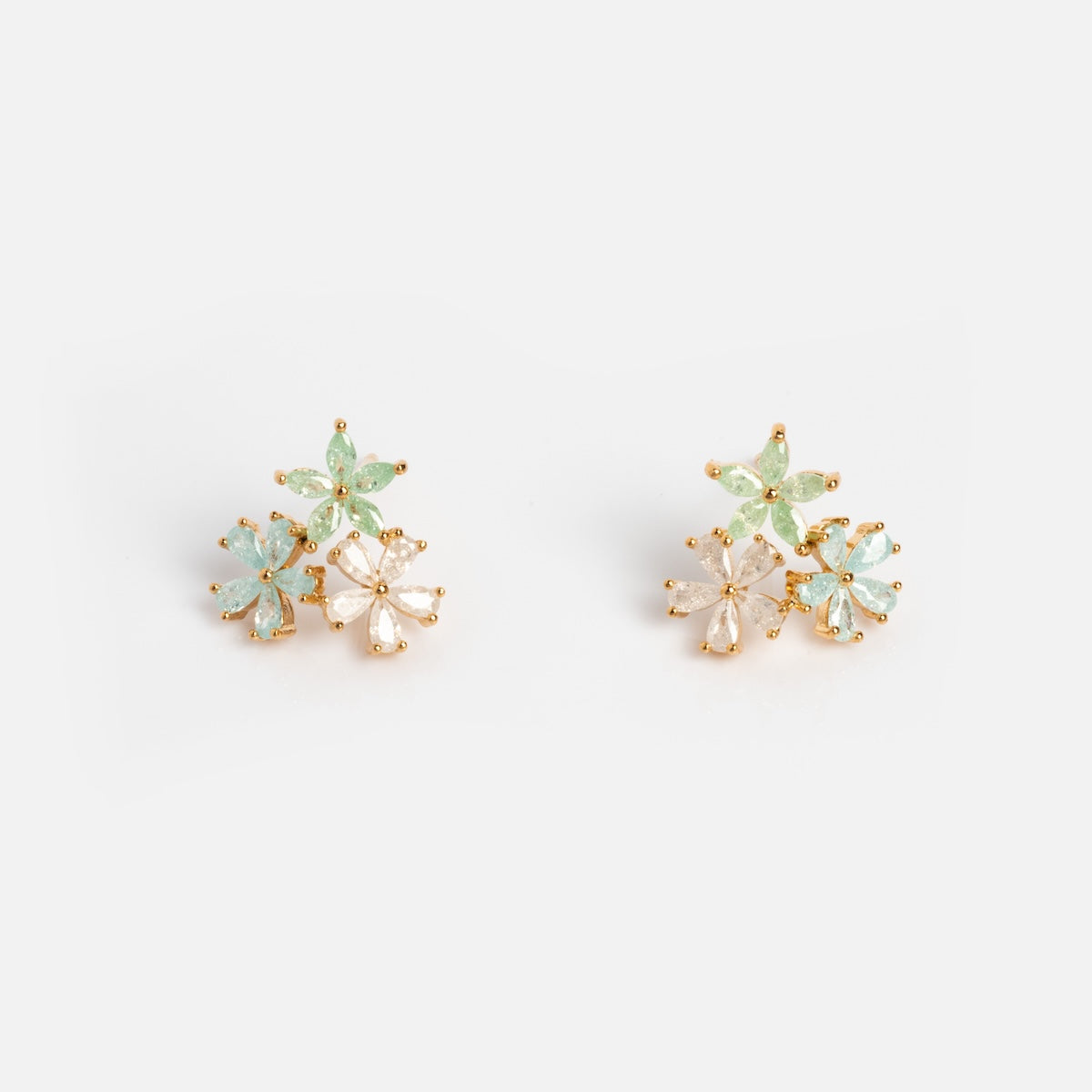 The Forget Me Not CZ Cluster Studs are 14k gold plated earrings featuring clusters of light green, pale blue, and clear petal-shaped stones delicately arranged as small flowers on a white background.