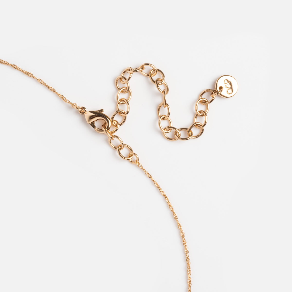 A close-up of the Muzi Pendant Necklace, 14k gold plated, featuring a gold chain with lobster clasp and adjustable extension lying on a white background. A small round tag is attached at the end of the extension chain.