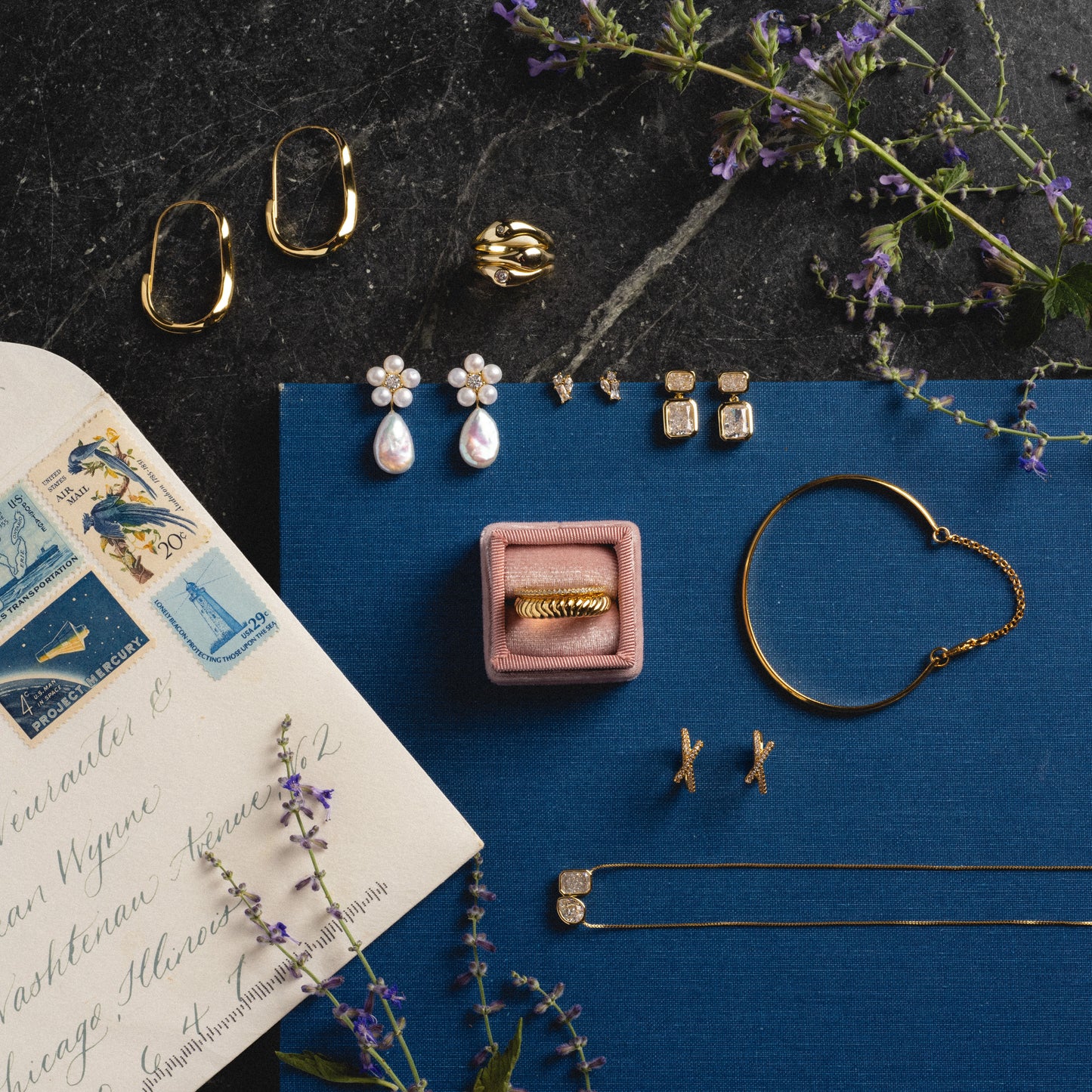 A flat lay featuring Flower Pearl Drop Earrings in 14k gold vermeil, alongside an envelope with vintage stamps, sprigs of purple flowers, and a blue textured background on a dark surface.