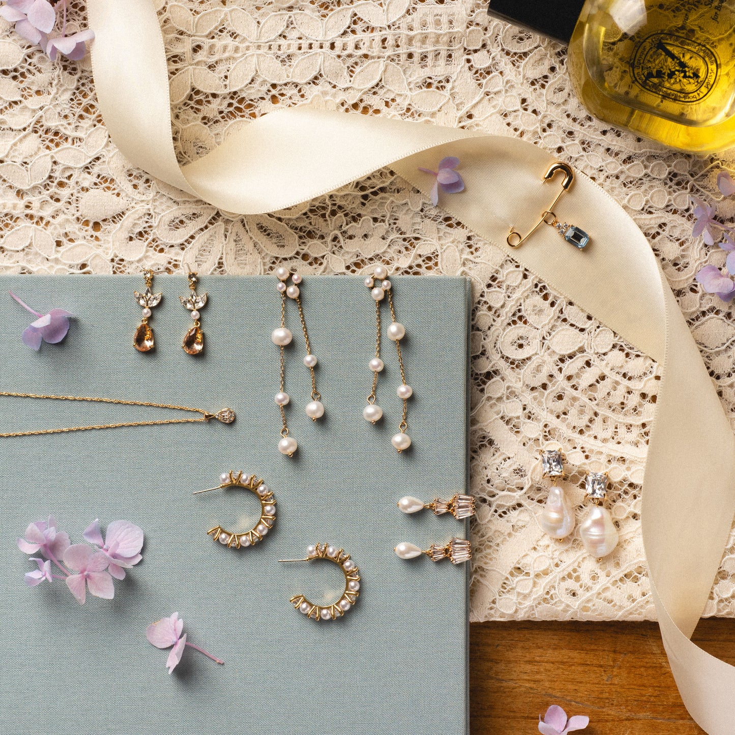A flat lay of elegant jewelry on lace and fabric, showcasing the Zeta Drop Earrings with freshwater pearls, a gold necklace, brooches, scattered purple flower petals, and a perfume bottle in the corner.