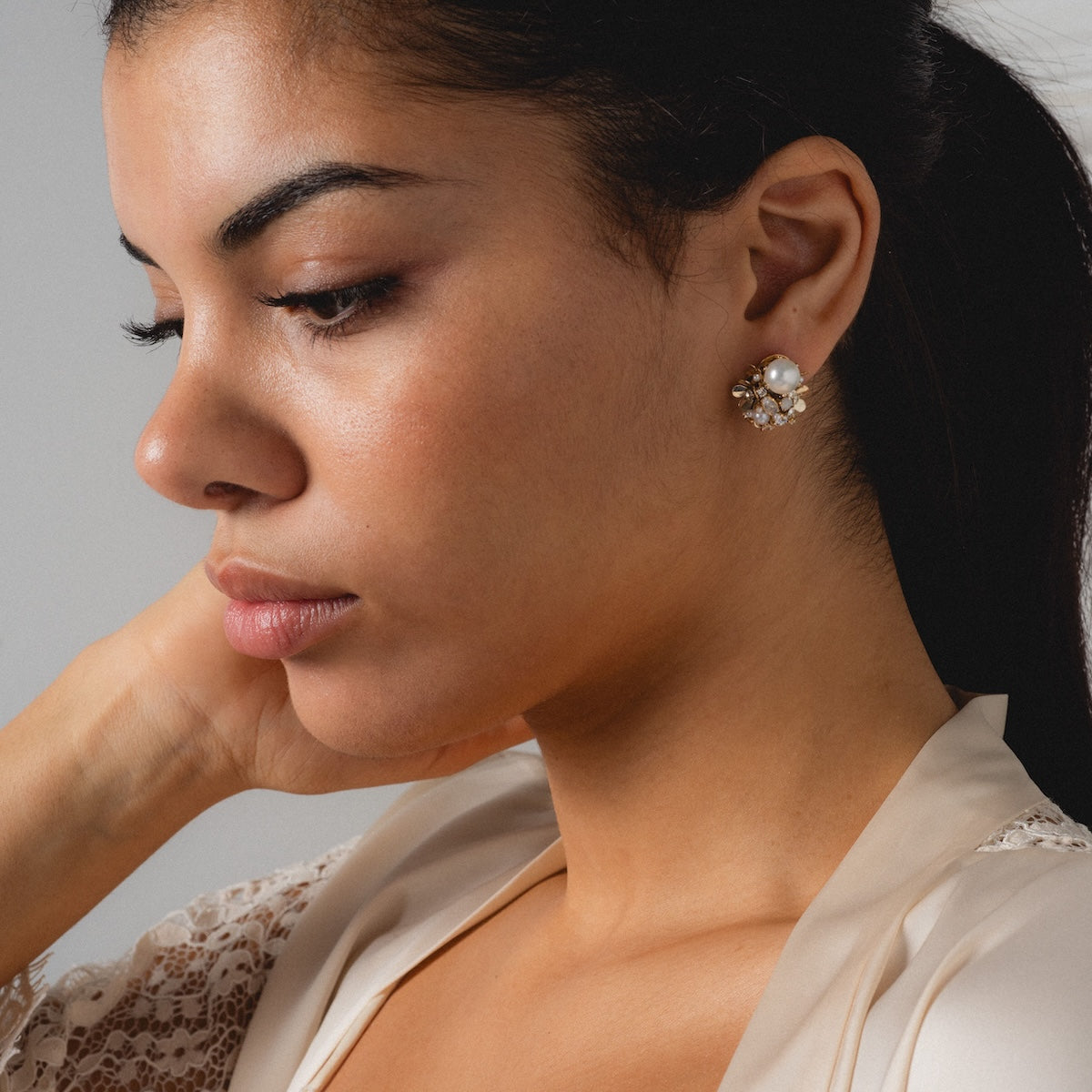 A woman with dark hair pulled back gazes downward thoughtfully. She wears a cream blouse with lace detail and the Luxe Pearl and Crystal Cluster Earrings, her hand gently resting on the back of her neck.