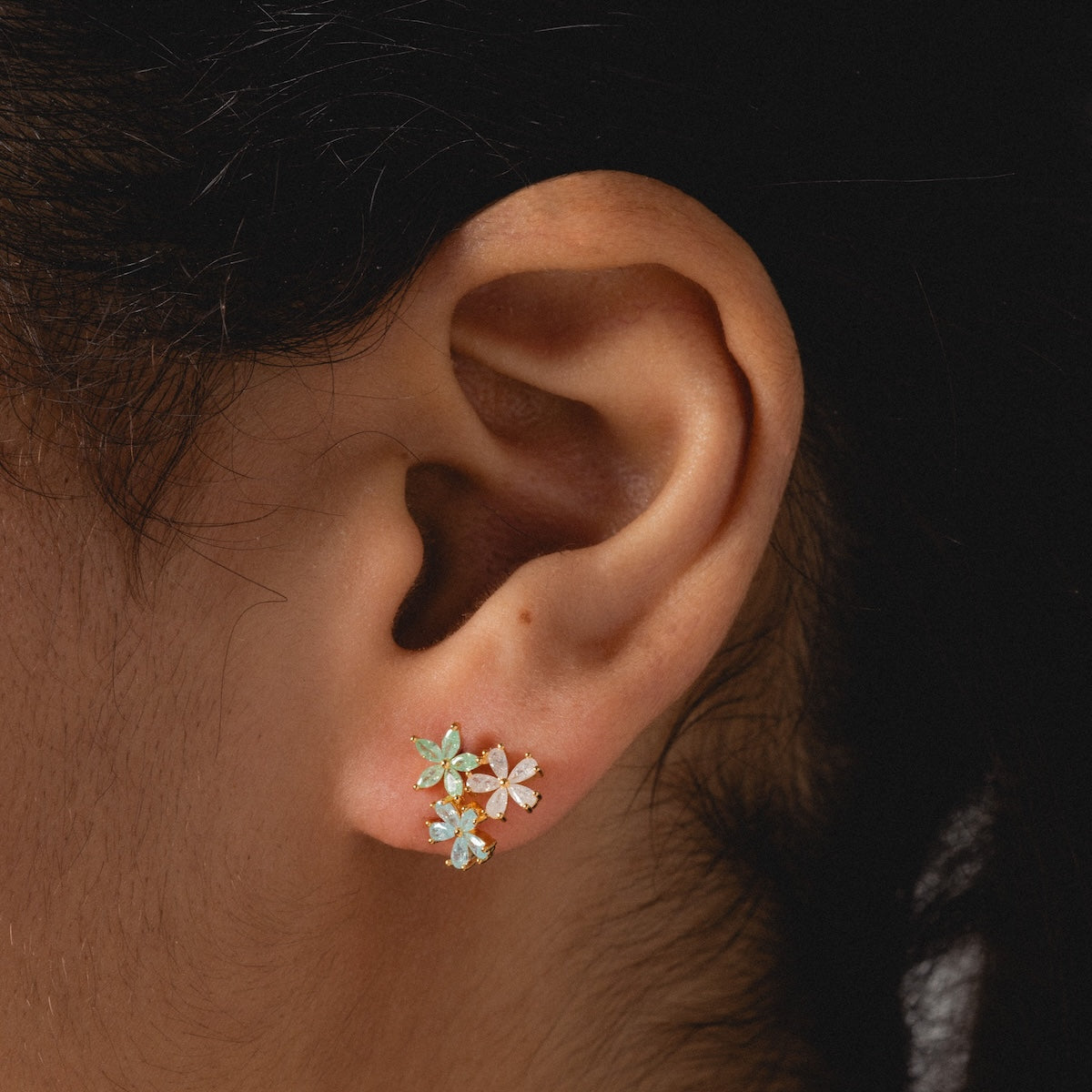 A close-up of a person’s ear wearing the Forget Me Not CZ Cluster Studs—romantic floral-shaped earrings with three pastel blue and white gemstones set in delicate 14k gold plating.