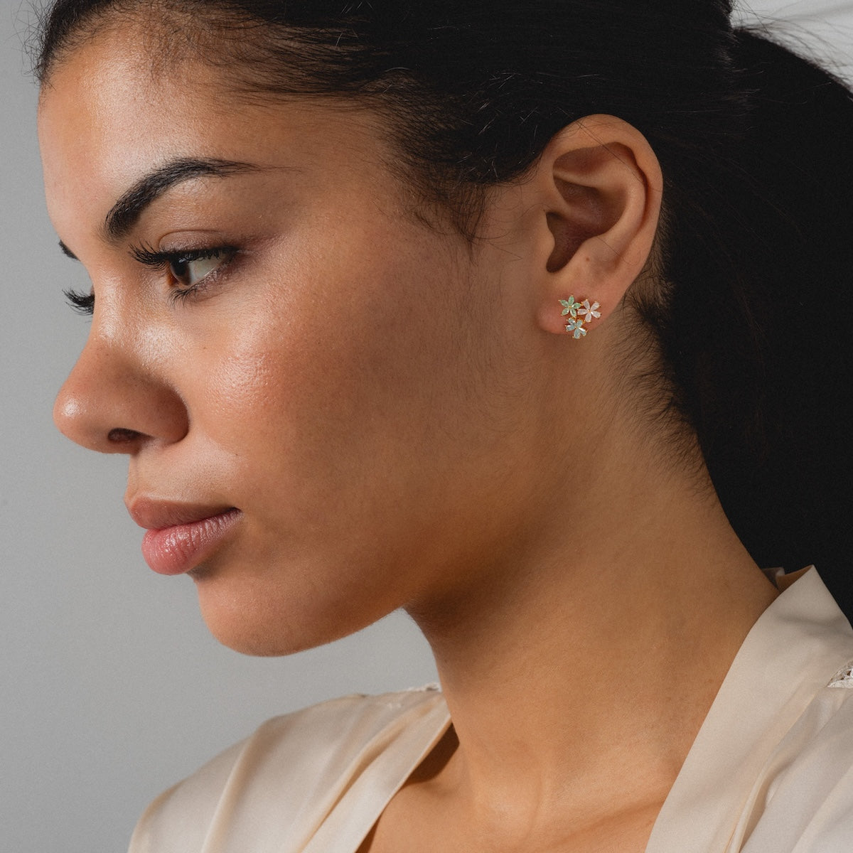 A woman with smooth skin and dark hair pulled back wears a cream blouse and the Forget Me Not CZ Cluster Studs—romantic 14k gold plated flower earrings—gazing thoughtfully to the side against a neutral background.