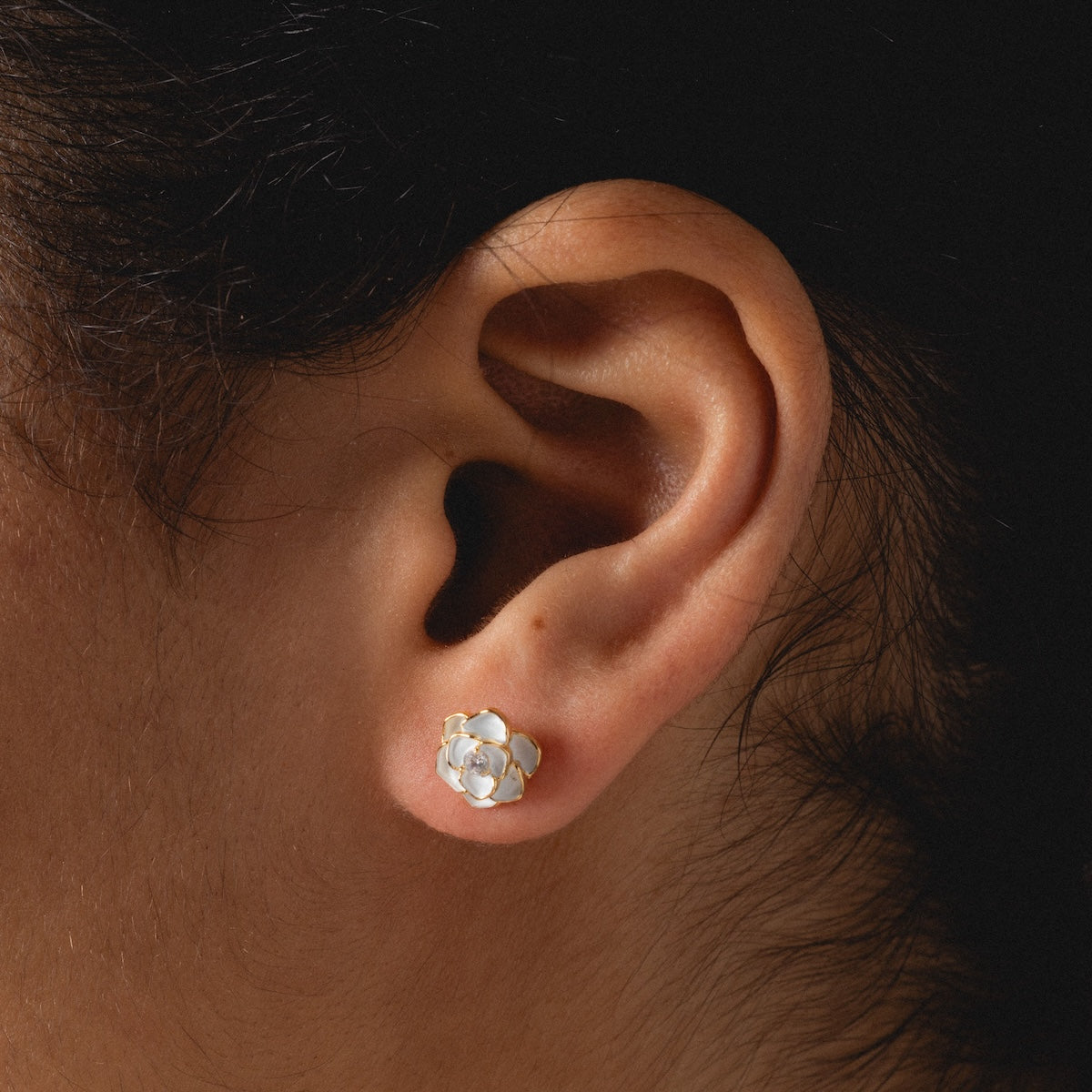 Close-up of a person’s ear adorned with Sparkling White Rose Studs—delicate enamel earrings shaped like flowers with white petals and a gold center. A softly lit, neutral background highlights the elegance of these floral studs.