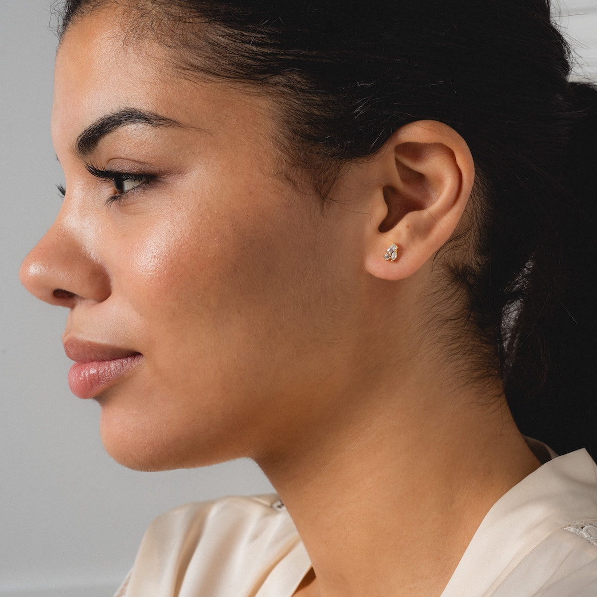 A woman with medium skin tone and dark hair in a ponytail is shown in profile, wearing Petite Pierre Studs—delicate yellow gold plated CZ crystal cluster earrings—and a cream top, looking slightly ahead with a neutral expression.