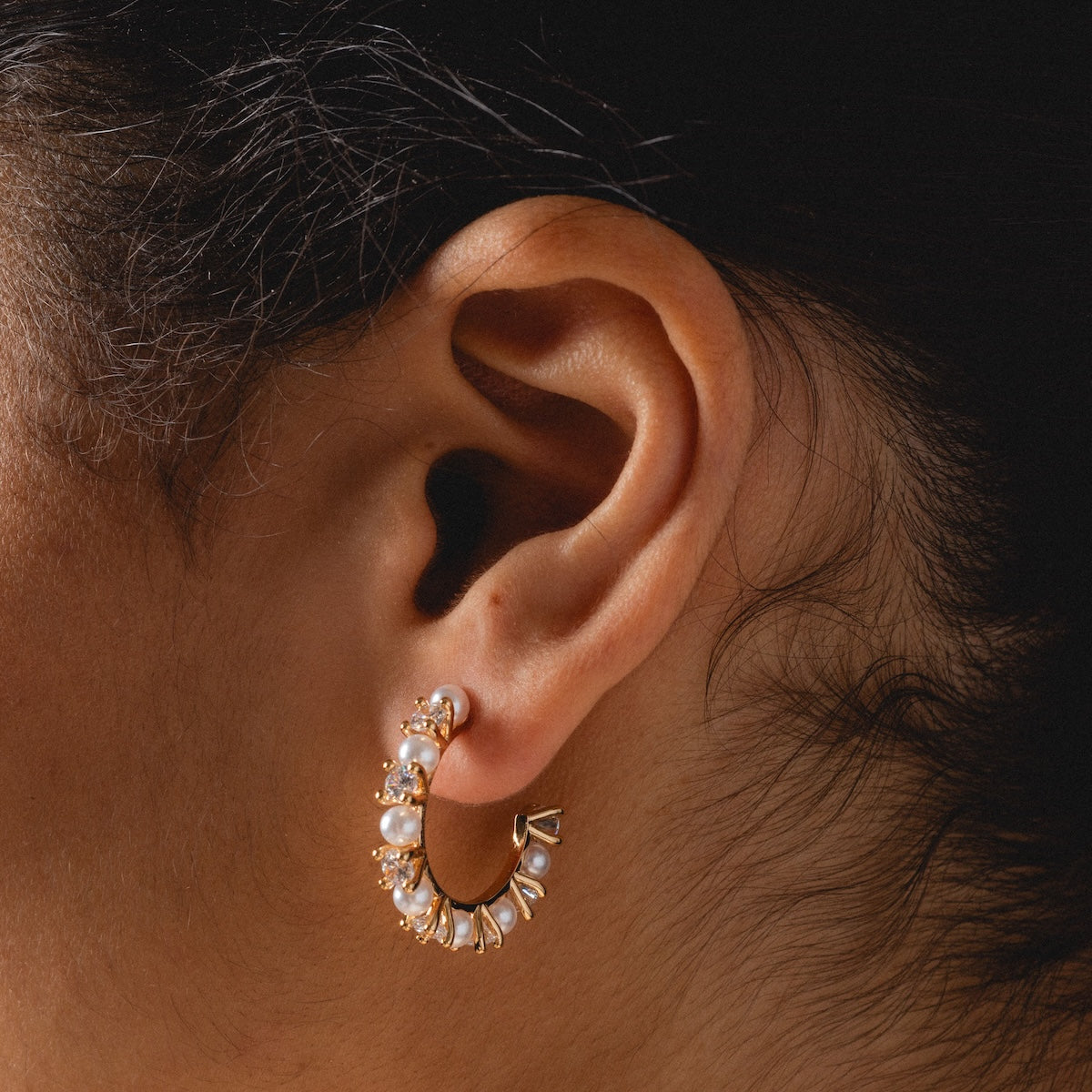 Close-up of a person’s ear wearing Prive Hoops—elegant gold plated earrings adorned with pearls and small gemstones—set against a neutral background.