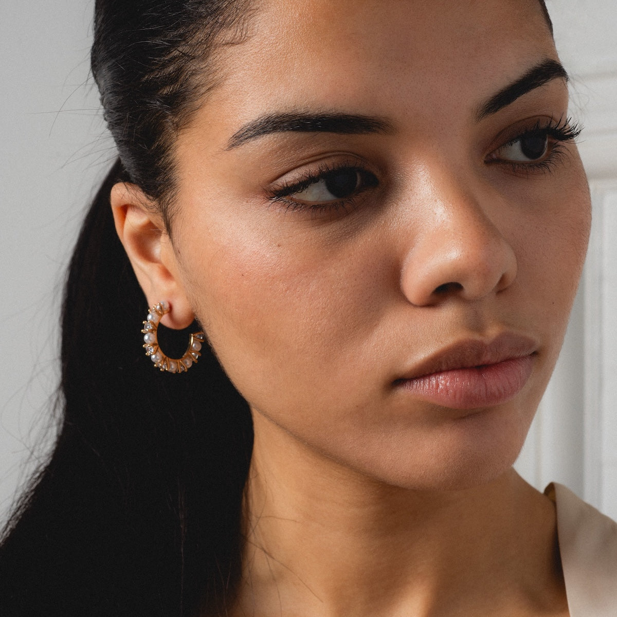 A woman with long dark hair in a ponytail wears the Prive Hoops—gold plated earrings with small crystal details. Photographed from the shoulder up against a neutral background, she appears thoughtful.