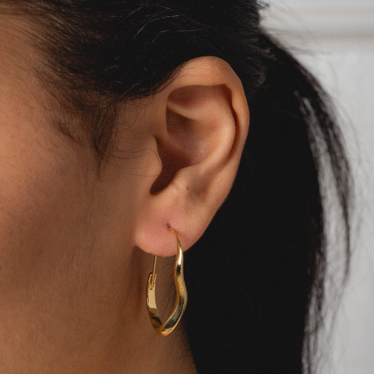 A close-up showcases the Mia Wave Hoops—elegant, shiny gold earrings—worn with dark hair pulled back to reveal the wearer’s ear and neck against a softly blurred background.