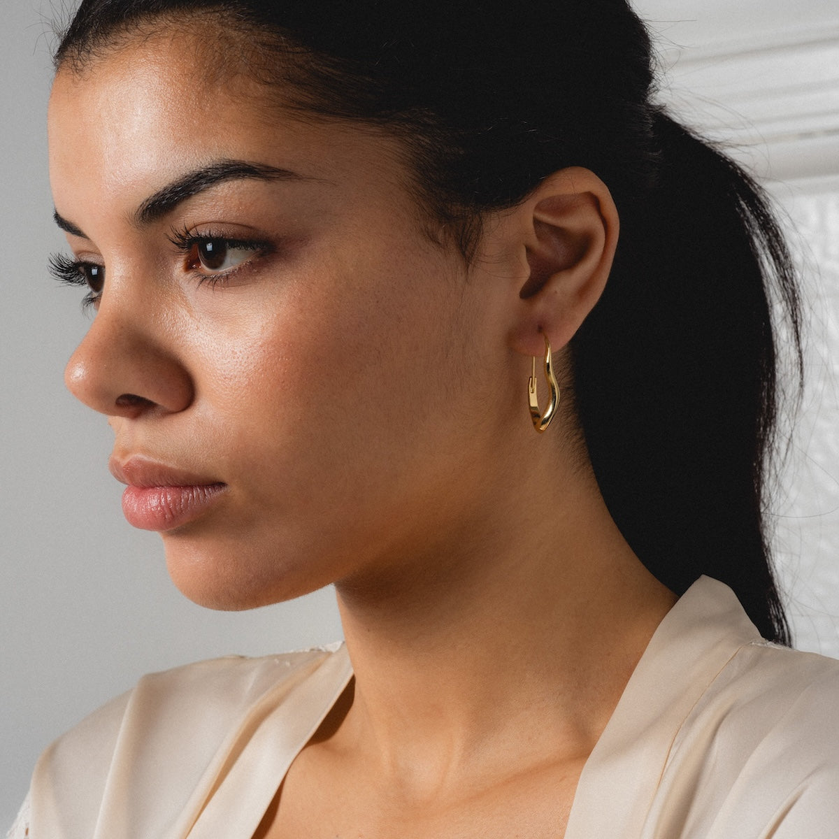 A woman with dark hair pulled back wears a cream top and the Mia Wave Hoops, gazing thoughtfully to the side against a neutral background.