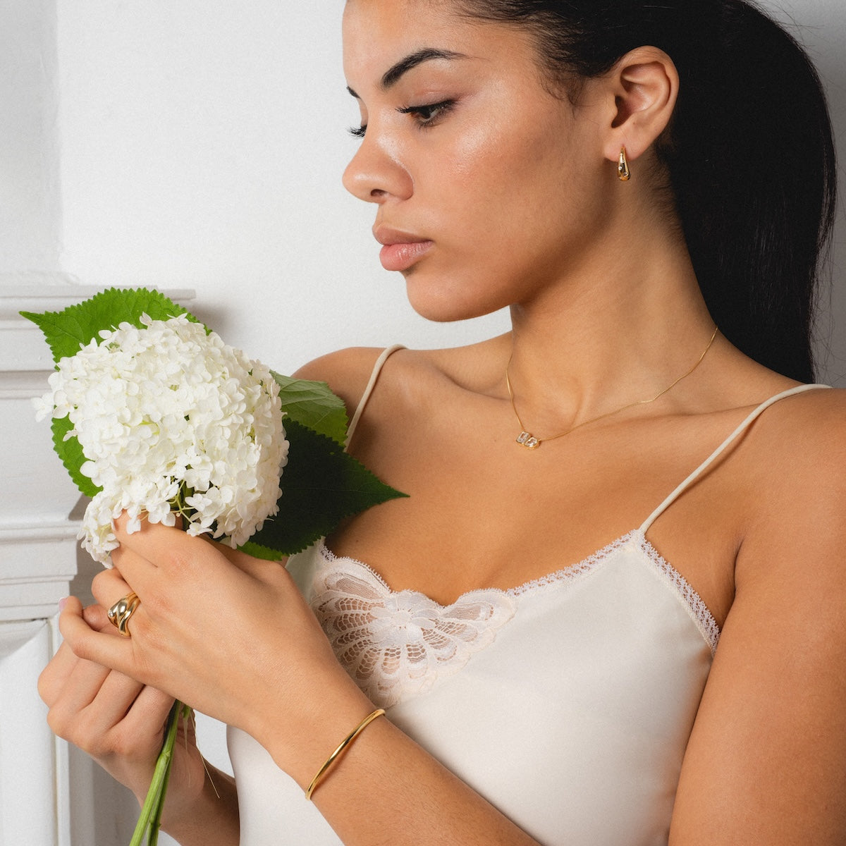 A woman in a cream-colored lace camisole holds white hydrangeas, wearing elegant gold jewelry including Shashi earrings and a 14k gold vermeil Baguette and Pear Cushion Pendant Necklace, gazing thoughtfully at the flowers.