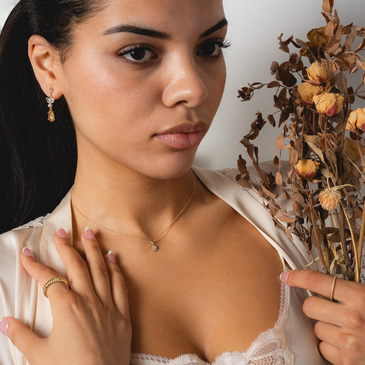 A woman wearing the Reagan Ring Set—14k yellow gold plated stacking bands—pairs them with a cream satin top, holding dried flowers and gazing calmly away from the camera.