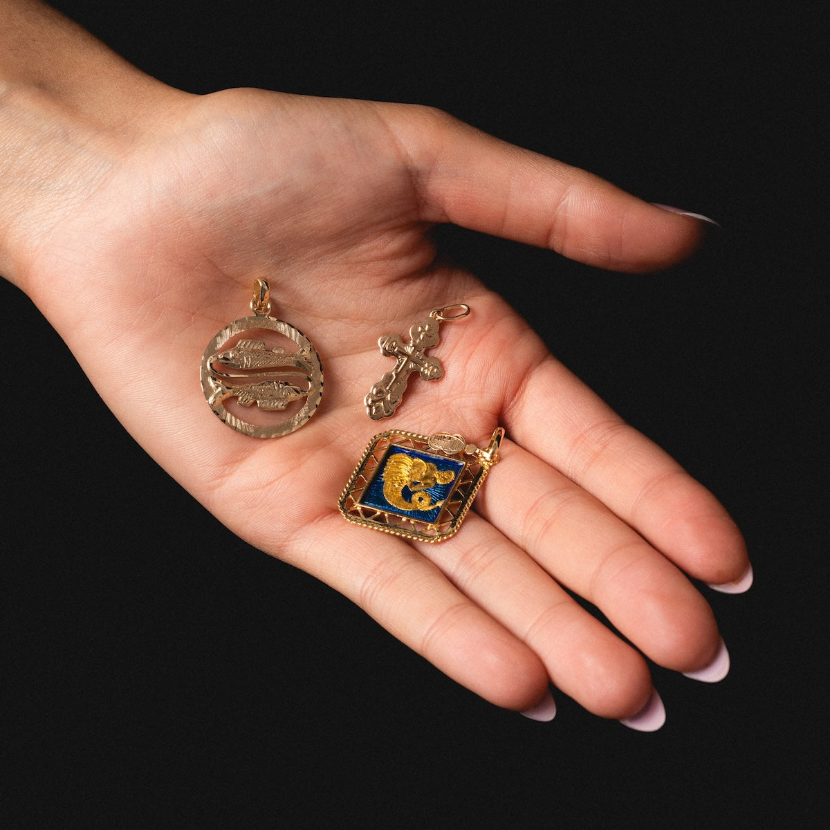 A hand with pale pink nails holds vintage jewelry: a round Vintage 14k Openwork Pisces Charm, a small ornate cross, and a square pendant featuring a blue background with a gold lion design.