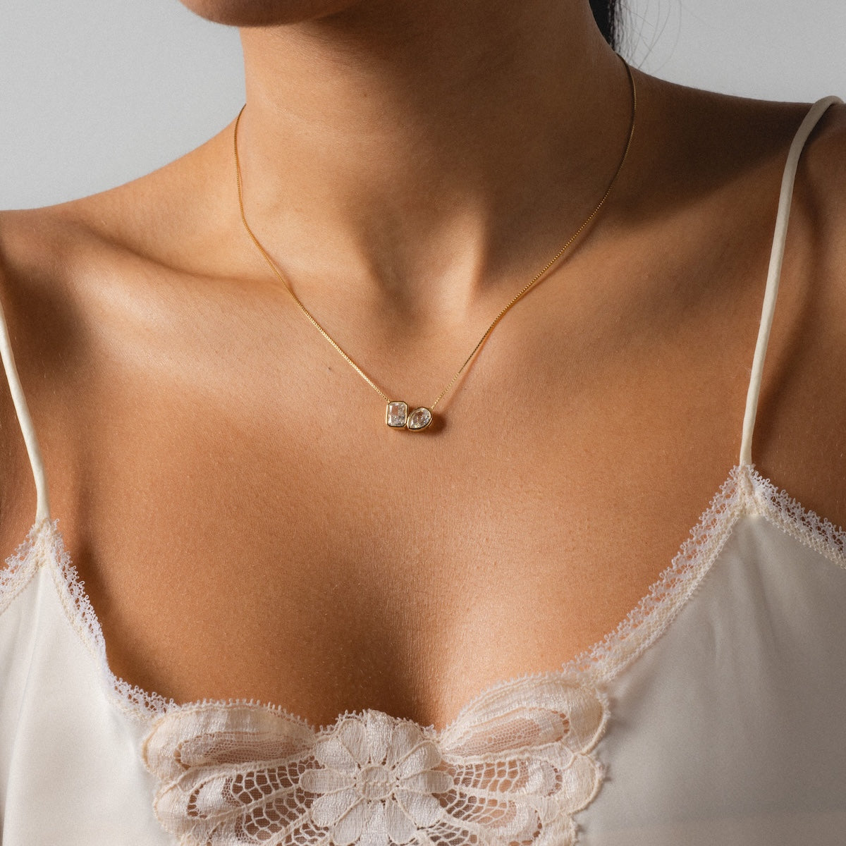 A woman wears the Baguette and Pear Cushion Pendant Necklace, paired with a cream-colored lace camisole featuring floral embroidery.