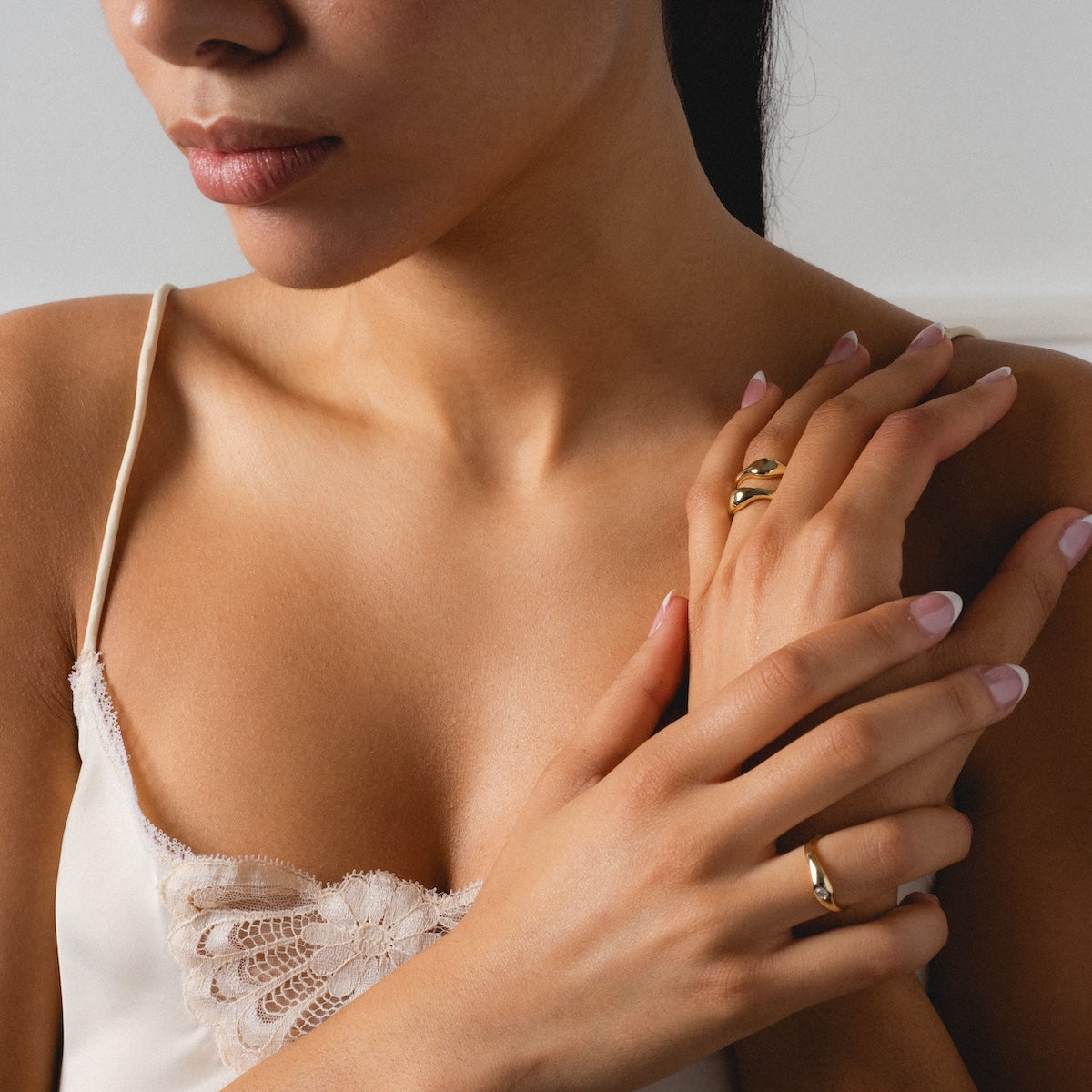 A woman in a white lace-trimmed camisole showcases the Delphine Solitaire Ring Set—wavy yellow gold rings on her manicured fingers—as she gently touches her collarbone. Only her lower face and upper torso are visible.