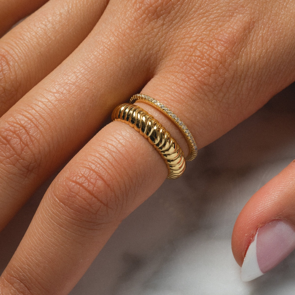 A close-up of a hand wearing the Reagan Ring Set—two 14k yellow gold plated rings, one thick and textured, one thin with small diamonds—stacked on the middle finger, paired with neatly manicured pale pink nails.