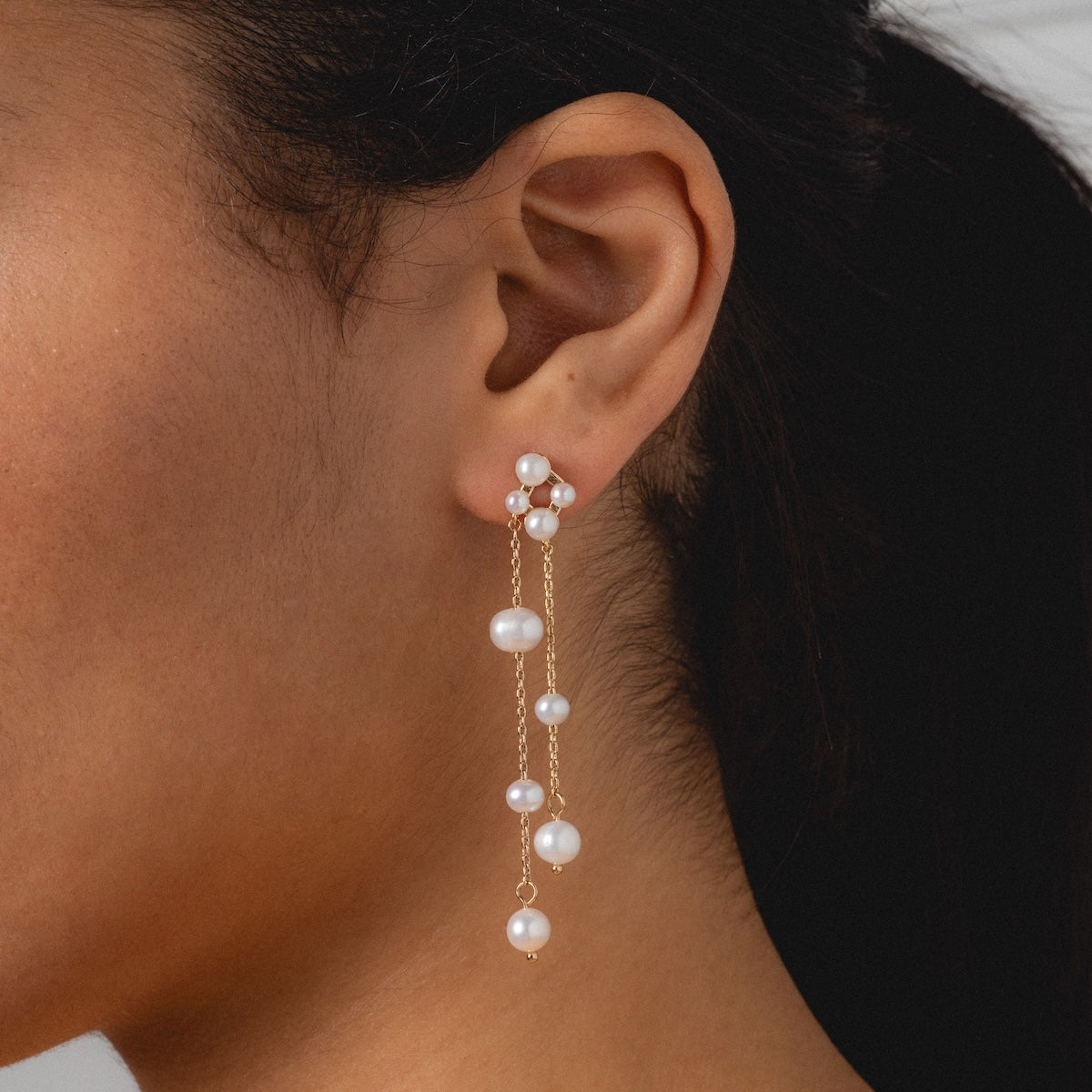 Close-up of a woman’s ear wearing Felicity Drop Earrings—gold plated with multiple white pearls along two delicate chains. Featuring both stud and drop design, these earrings are perfect for showcasing elegant freshwater pearl style.