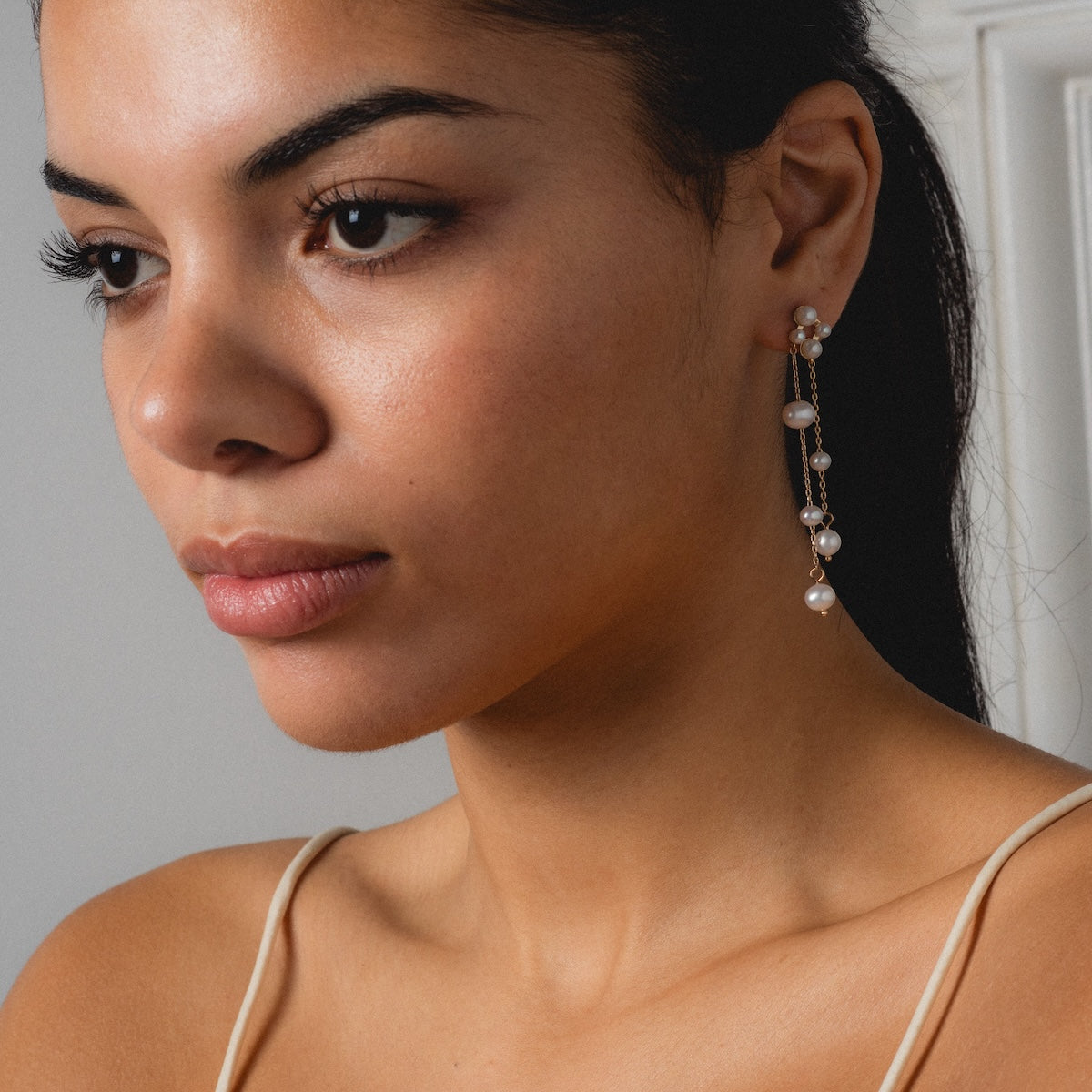 A woman with smooth, medium-toned skin and dark hair wears the Felicity Drop Earrings—long, elegant gold-plated earrings with pearls. She is dressed in a beige spaghetti-strap top, gazing thoughtfully to the side against a light background.