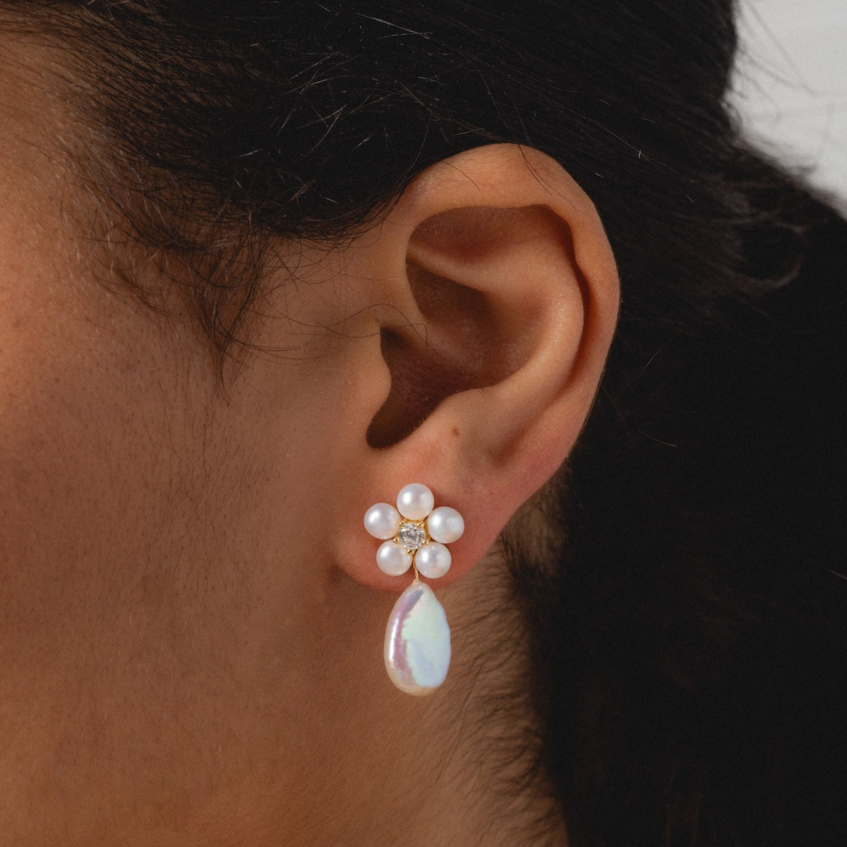 Close-up of a person with dark hair pulled back, wearing Flower Pearl Drop Earrings—14k gold vermeil earrings with a flower design made of five round freshwater pearls, a central gem, and a teardrop-shaped pearl drop.