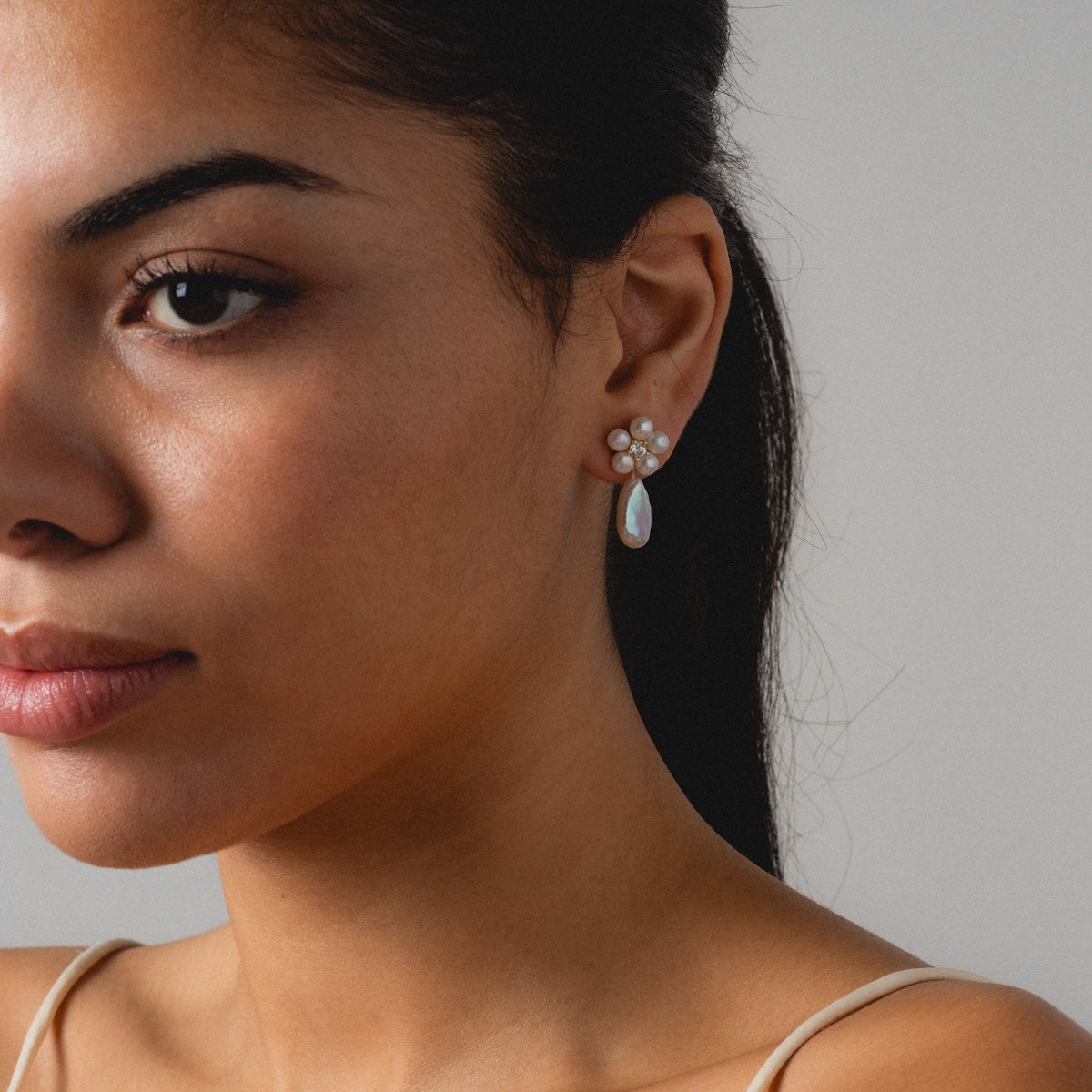 A woman with smooth skin and dark hair wears Flower Pearl Drop Earrings, crafted in 14k gold vermeil and freshwater pearls, in a close-up portrait against a neutral background. She gazes softly ahead.