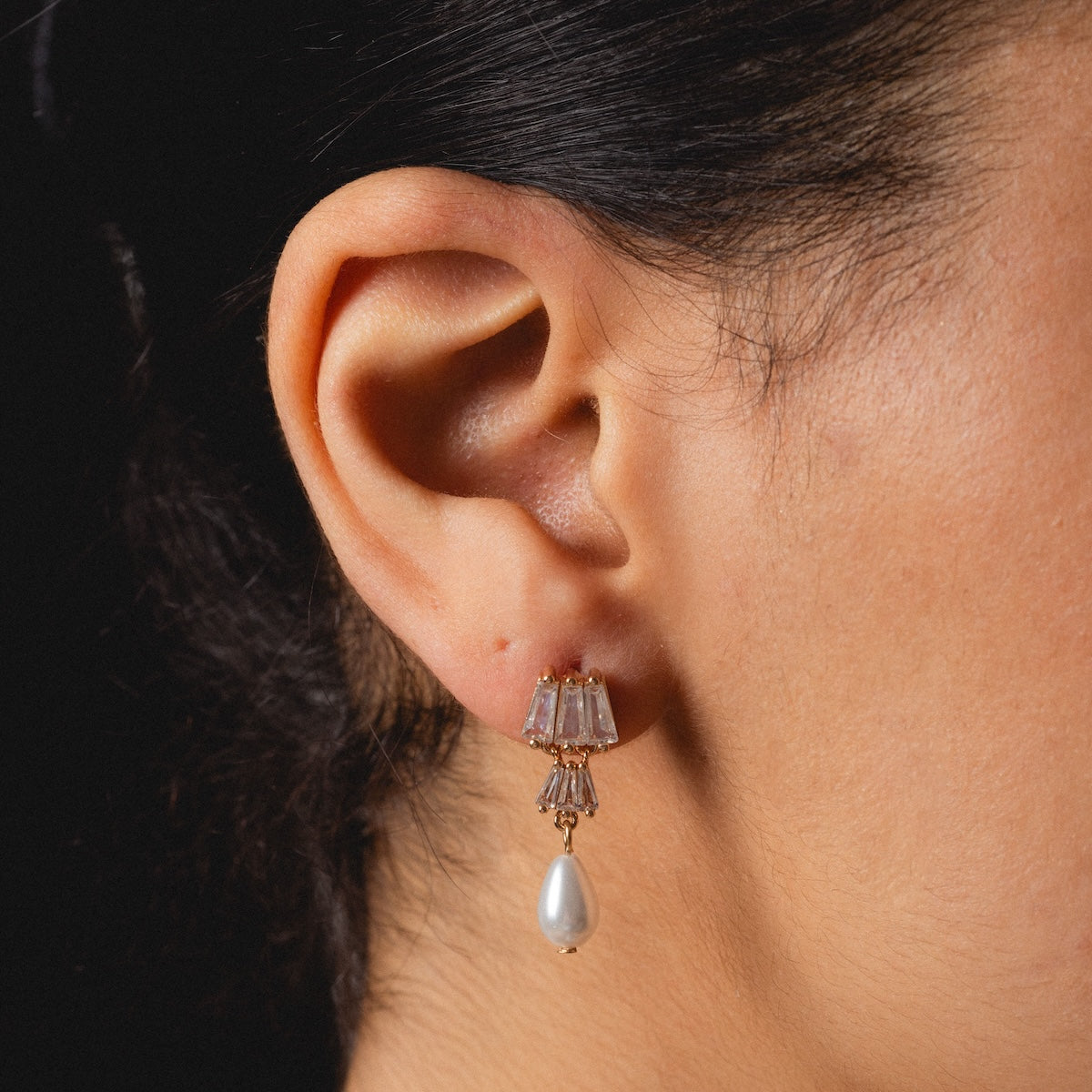 Close-up of a persons ear wearing Rue Drop Earrings, featuring clear gemstones and a dangling white pearl. The dark background highlights the elegant jewelry and the persons skin.