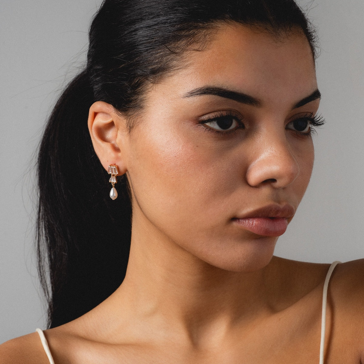 A woman with long dark hair pulled back wears the elegant Rue Drop Earrings. She has smooth, natural makeup and looks slightly to the side against a plain, light background.