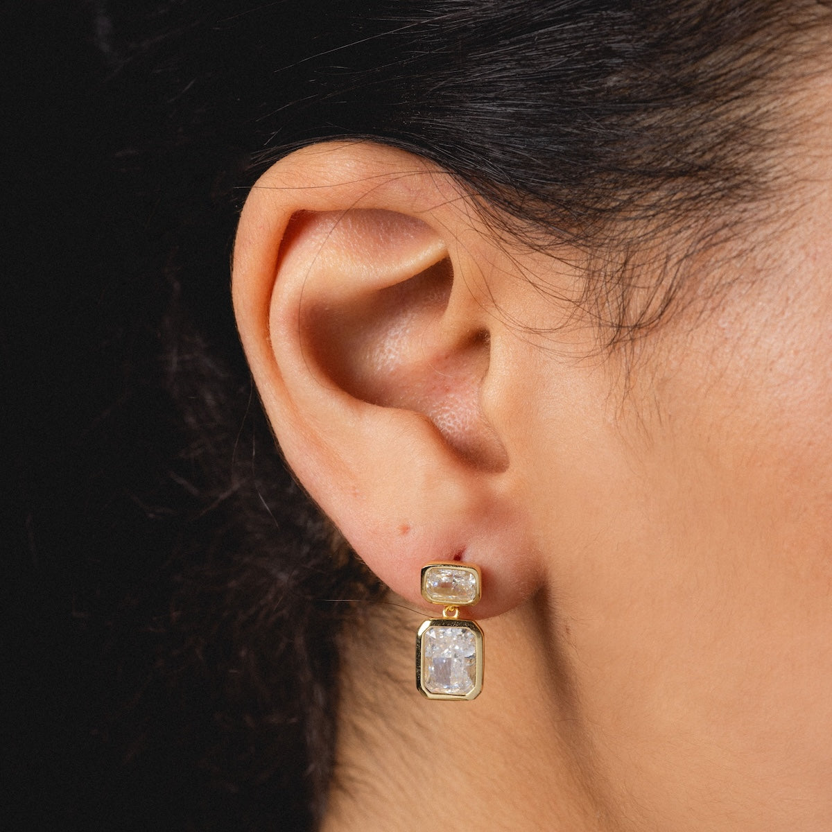 Close-up of a womans ear wearing Emerald Baguette Drop Earrings in 14k gold vermeil with two clear rectangular gemstones, one small and one large, set vertically. Her hair is pulled back against a black background.