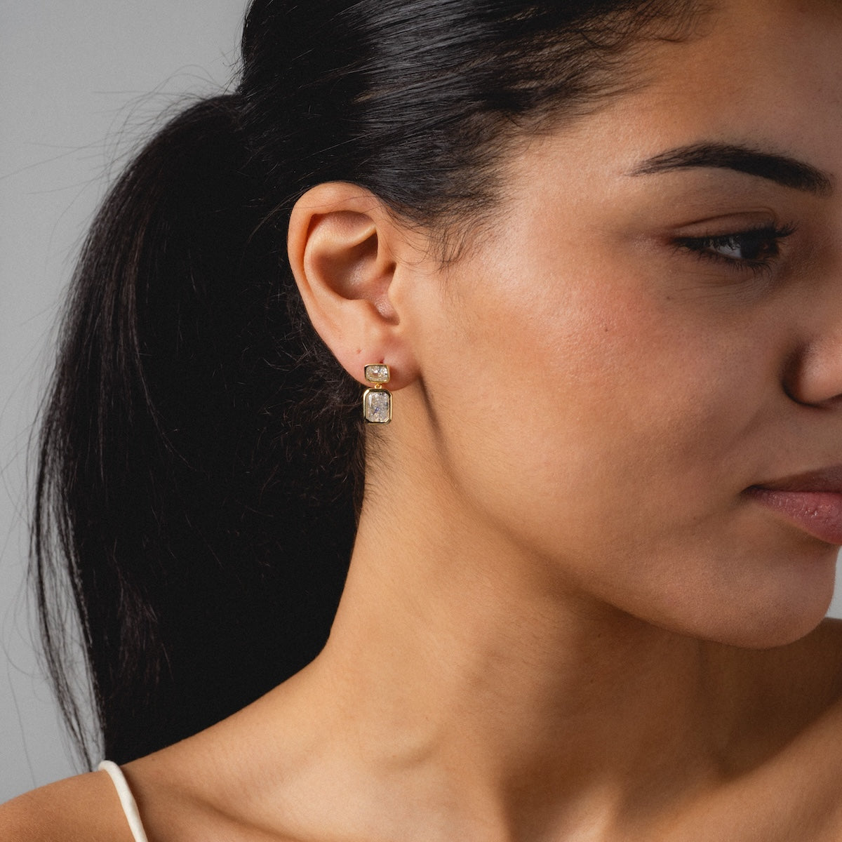 A woman with long dark hair in a ponytail, seen from the side against a neutral background, wears a sleeveless top and Emerald Baguette Drop Earrings.