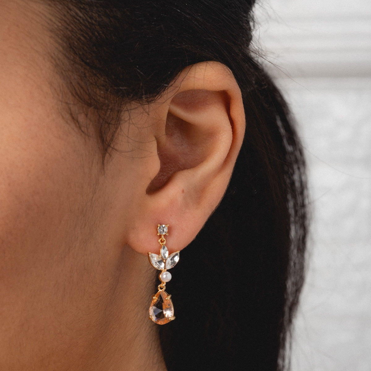 Close-up of a womans ear wearing Mini Rosalind Drop Earrings—gold plated with clear gemstones and a teardrop-shaped crystal—set against a neutral background.