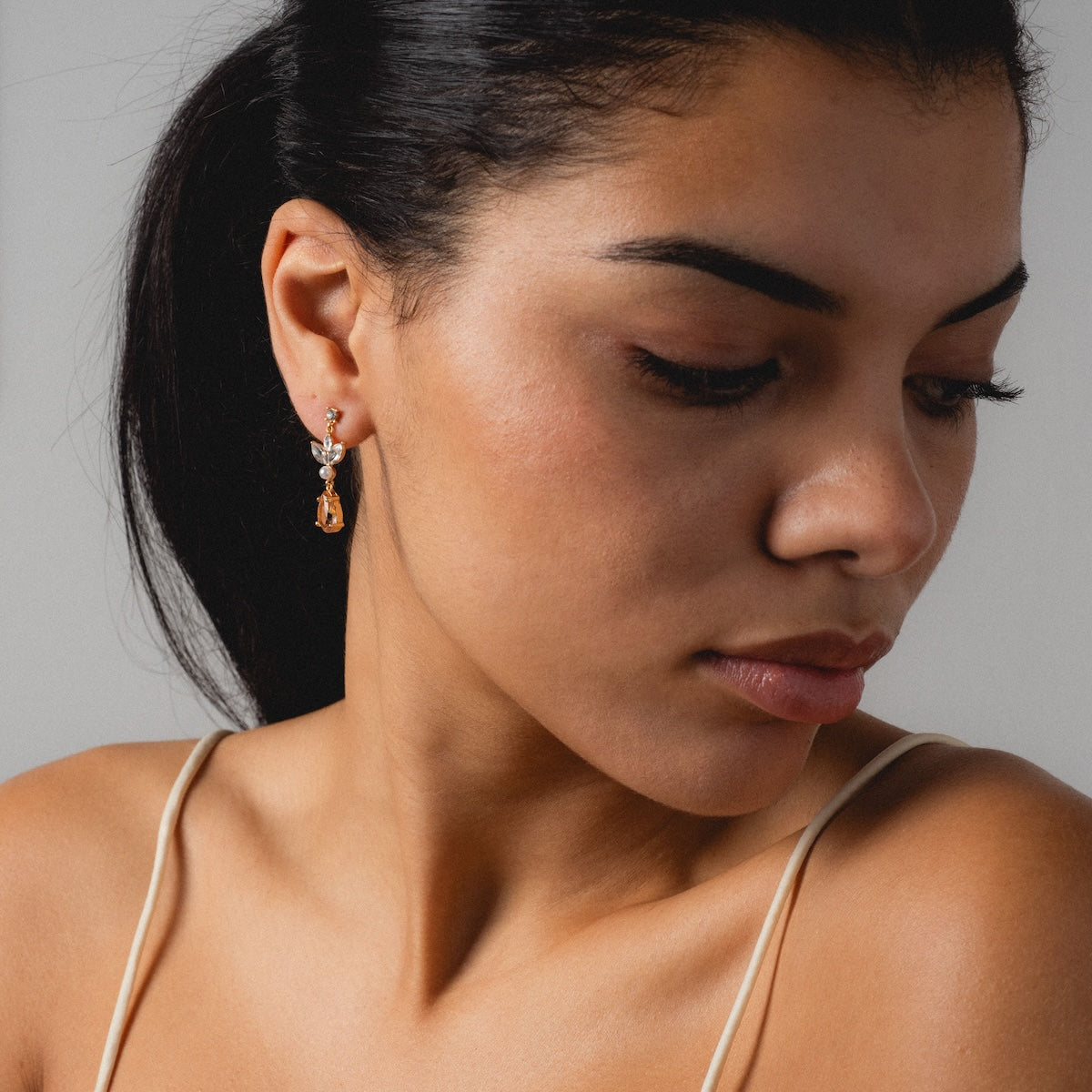 A woman with smooth skin and dark hair looks down, wearing the Mini Rosalind Drop Earrings and a thin strap top, set against a plain light background.
