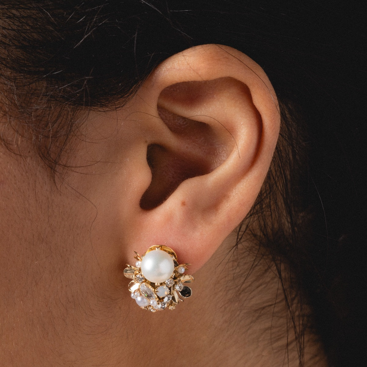 Close-up of a persons ear wearing Luxe Pearl and Crystal Cluster Earrings—gold plated with a large central pearl, smaller white pearls, and clear crystals in a floral cluster, ideal for lovers of pearl cluster earrings.