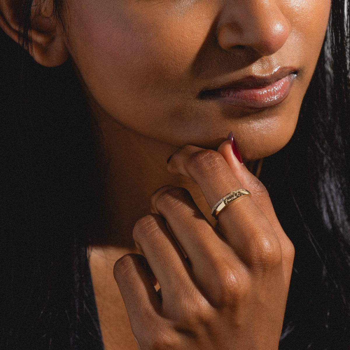 A close-up of a woman with medium brown skin and dark hair gently touches her chin, showcasing the Solid Gold Tiny Fuck Sapphire Stacking Ring Set. Soft lighting highlights the gold ring’s engraved design on her hand.